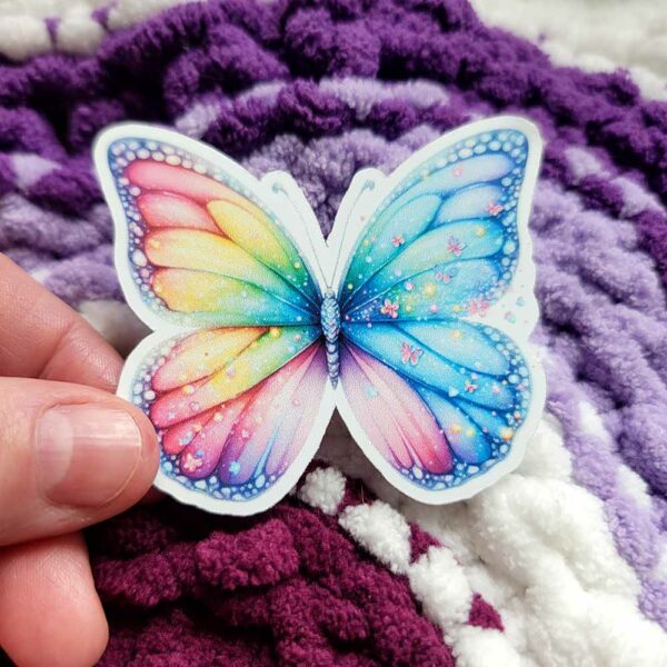 A vibrant die-cut vinyl sticker of a rainbow-colored butterfly with glittery wings, adorned with tiny butterfly motifs and sparkling accents.