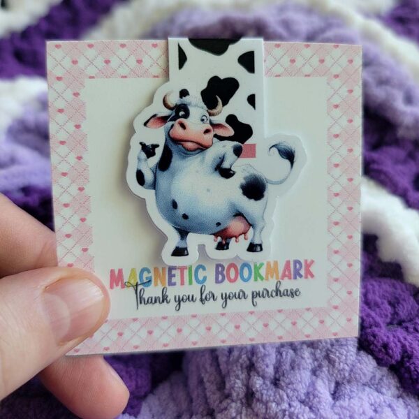 Cartoon-style cow with a playful expression, black and white spots, and a pink snout, used as a magnetic bookmark.