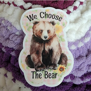 A vinyl sticker featuring "Bruno" the Bear with brown and white fur, surrounded by sunflowers and a pink gingham background. Text reads "We Choose The Bear."