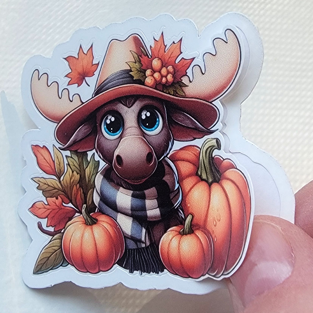 Vinyl sticker of a cute moose wearing a hat and scarf, surrounded by pumpkins and autumn leaves. Demonstration of removing sticker from backing with easy peel stickers